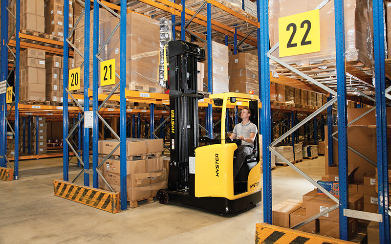 Reach truck lifting a pallet in a high-density warehouse with tall racking and narrow aisles.