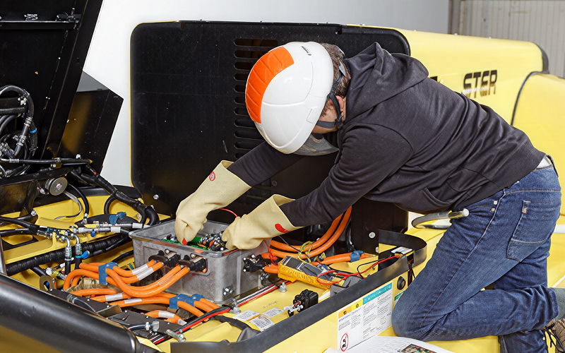Southeast Asian warehouse technician in a safety vest and hard hat inspecting a forklift’s open battery compartment in a marked charging bay inside a large warehouse.